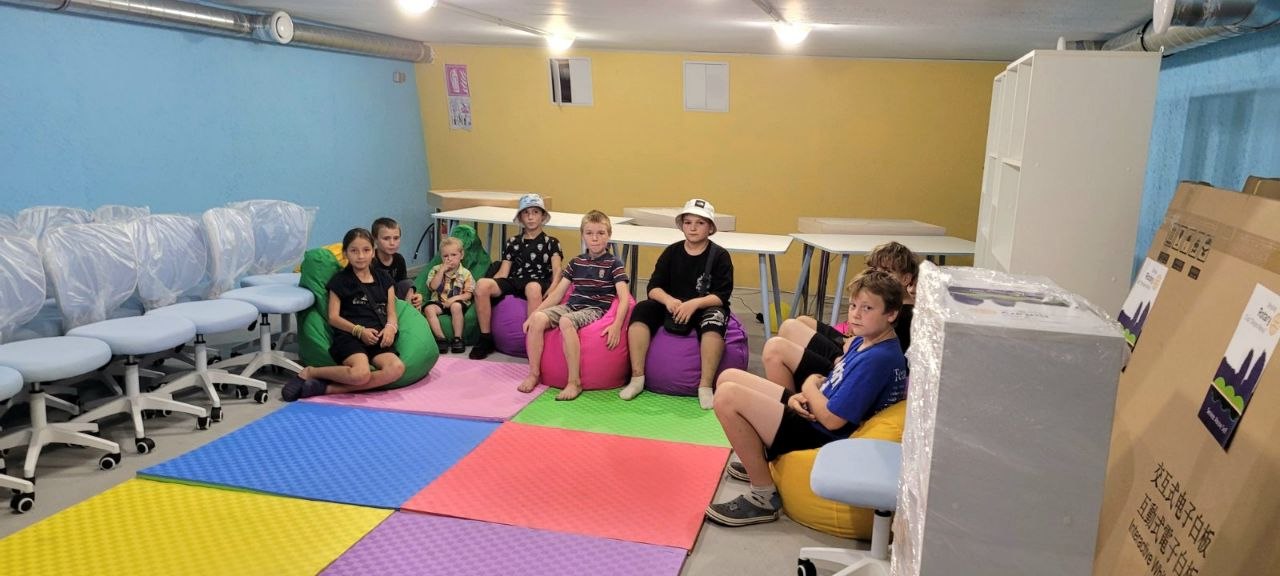 Wider view of students on bean bags in the newly outfitted underground room, with soft floor mats and incoming equipment visible, August 2026.