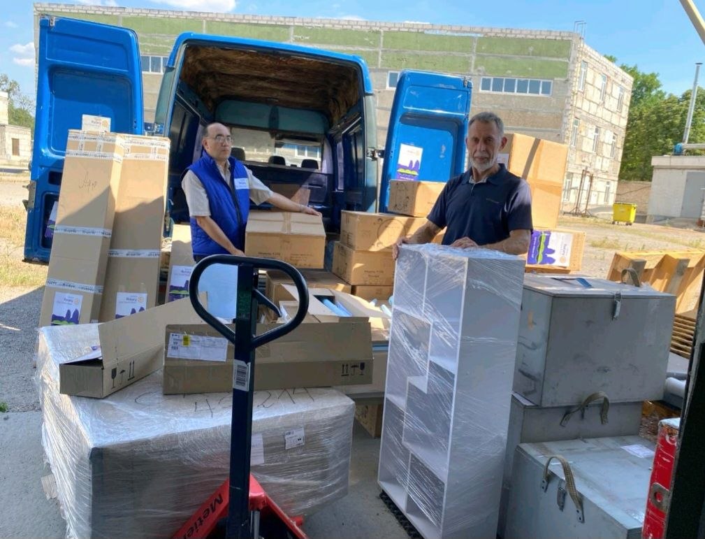 Two Rotarians standing behind a van full of supplies for Vyvodove, boxes marked Ukraine Rotary Club Dnipro Novyi, August 2026.