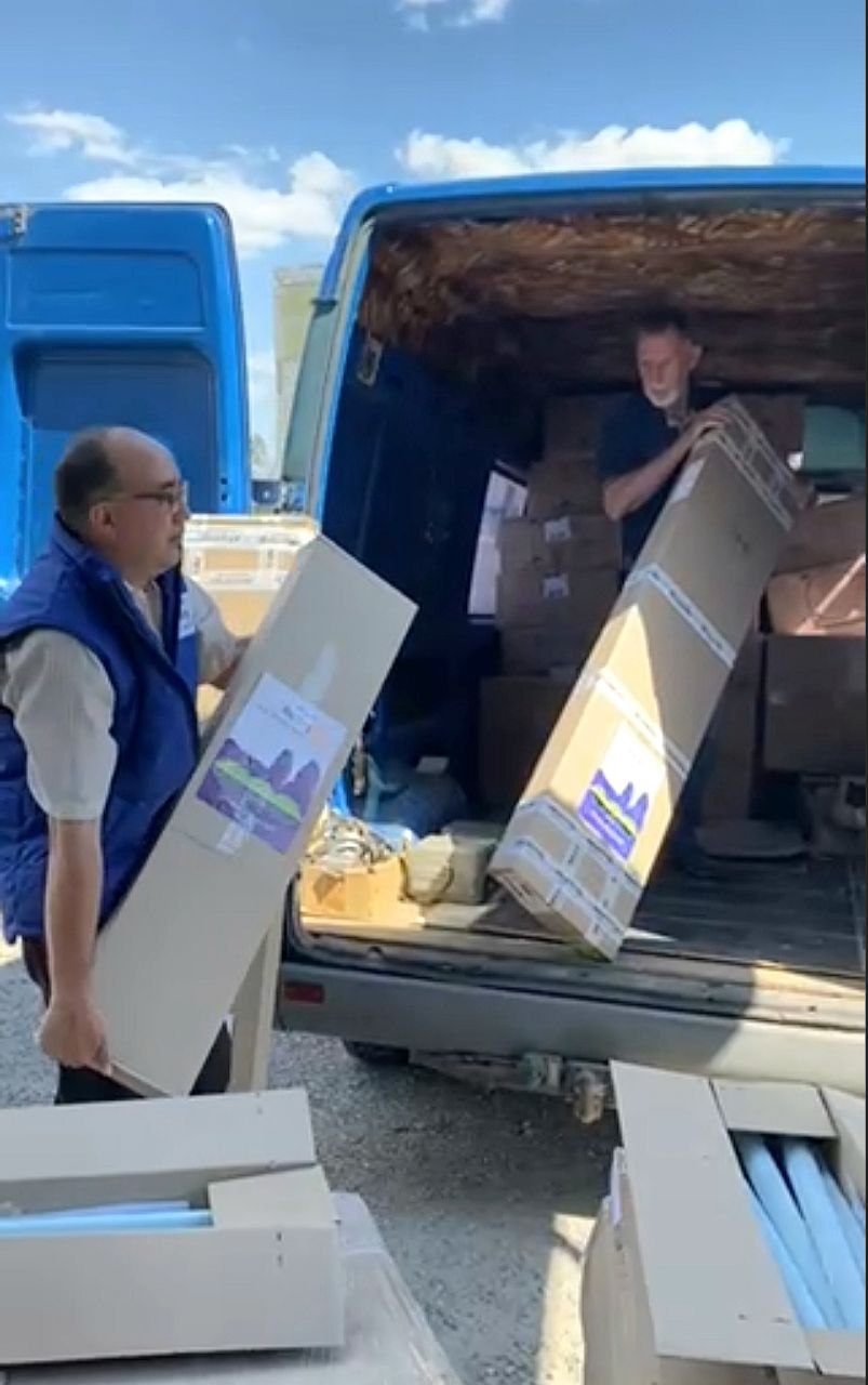 A Rotarian in a blue vest carrying a labeled Rotary Club of Dnipro Novyi box during the August 2026 delivery to Vyvodove.