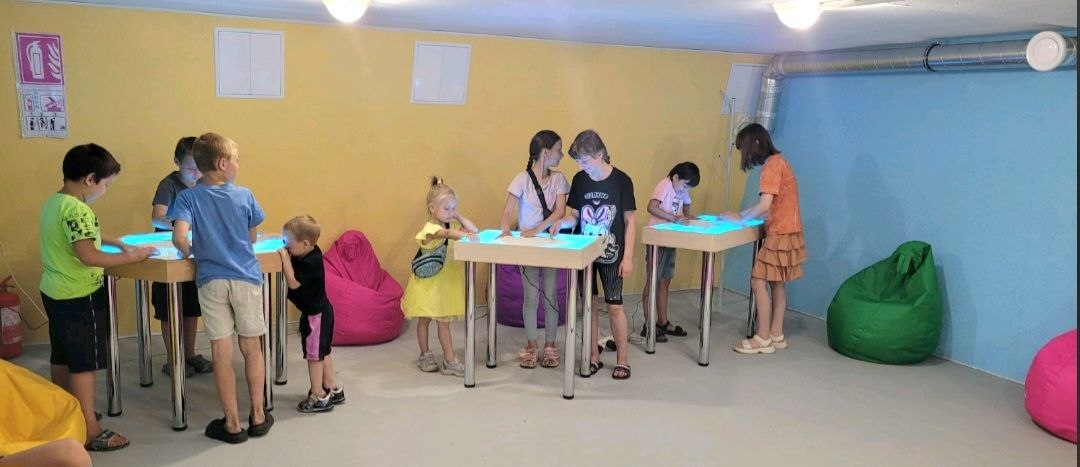 Children drawing on three backlit sand-and-light therapy tables in the completed underground Peace & Mental Health Resource Center at Vyvodove, August 2026.